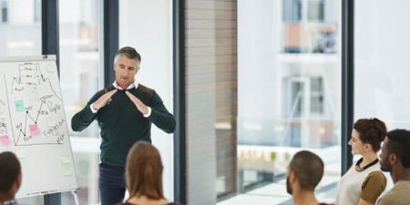 Un homme debout devant un paperboard en pleine explication face à un public d'adultes