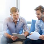 Deux hommes, l'un jeune l'autre senior, discutent, avec des documents en main